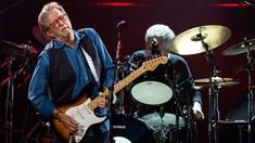 an older man playing guitar on stage with other people in the background and one person standing next to him