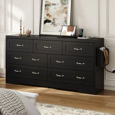 a black dresser with gold handles in a white room next to a painting and lamp