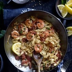 a pan filled with pasta and shrimp on top of a blue cloth next to lemon wedges