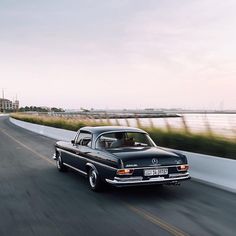 an old black car driving down the road next to a body of water with buildings in the background