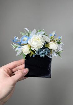 a hand holding a black box with white and blue flowers in it on a gray background