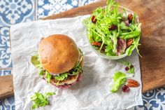 a sandwich with lettuce and other vegetables on a cutting board next to a bowl of salad