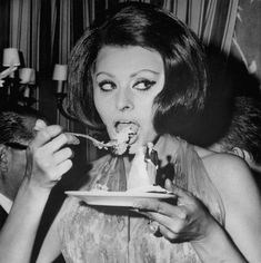 a woman eating cake from a plate with a fork in her mouth while another man looks on