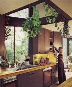 a woman is in the kitchen with plants hanging from the ceiling and she is washing dishes