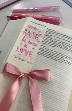 an open book with writing on it and a pink ribbon tied around the page, next to two pens