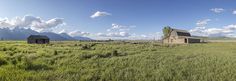 an old barn sits in the middle of a grassy field with mountains in the background