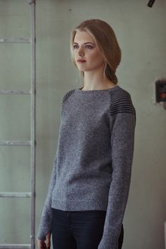 a woman standing in front of a ladder wearing a gray sweater and black pants with her hands in her pockets