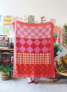 a woman holding up a pink and red quilt in front of a wall with pictures on it