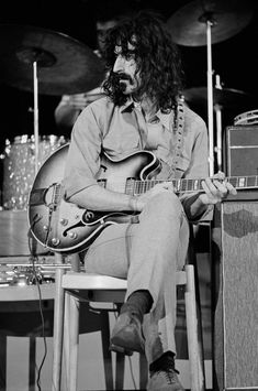 a man with long hair sitting on a chair playing an electric guitar in front of two amps