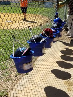 blue buckets filled with baseball equipment sitting on the side of a fenced in field