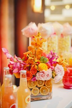 an arrangement of flowers and oranges in vases on a table with soda bottles