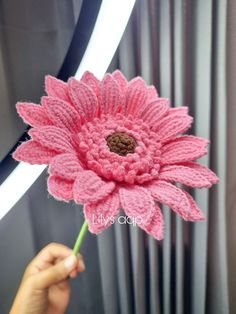 a person holding a pink crocheted flower in front of a window sill