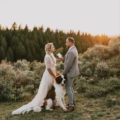 a bride and groom standing next to their dog in the grass with trees in the background