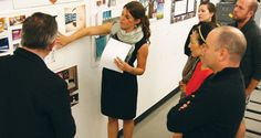 a group of people standing in front of a white wall with pictures on it and one person pointing at something