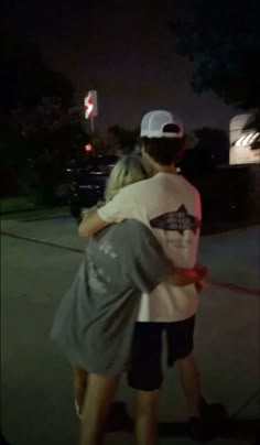 two people hugging each other while standing on the sidewalk at night with their arms around each other