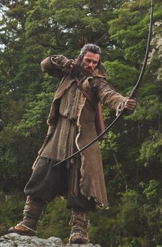 a man with long hair holding a bow and arrow while standing in front of trees