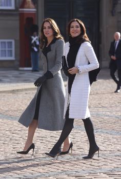 two women in coats and black shoes walking down a cobblestone street with buildings behind them