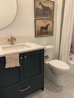 a white toilet sitting next to a bath tub under a bathroom mirror with two pictures above it