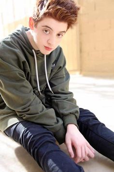 a boy sitting on the floor with his legs crossed wearing black pants and a green hoodie