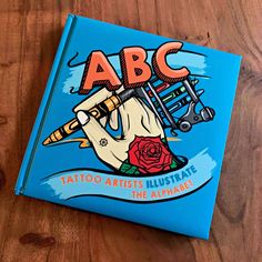 an abc tattoo artist's illustrated book sits on a wooden table next to a pen