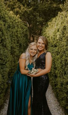 two women standing next to each other in front of some bushes and trees with their arms around each other