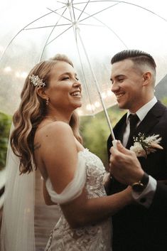 a bride and groom standing under an umbrella smiling at each other while holding their wedding bouquet