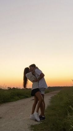 a man and woman kissing on the side of a dirt road in front of a sunset