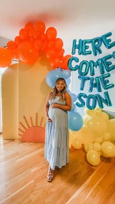 a pregnant woman standing in front of balloons and the words here comes the son on it