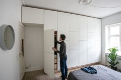 a man standing next to a bed in a room with white cupboards and drawers