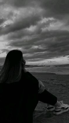 a woman standing on the beach looking out at the ocean with clouds in the sky