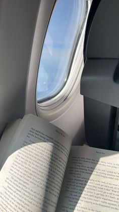 an open book sitting on top of an airplane seat next to a window that looks out onto the sky