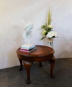 a wooden table with a vase and flowers on it next to a white busturine