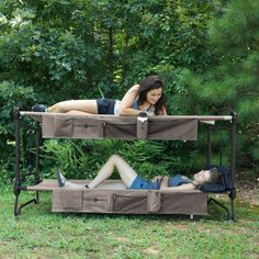 two women laying on top of bunk beds in the grass next to some trees and bushes