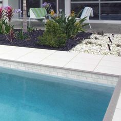 an empty swimming pool in front of a house with lawn chairs and flowers around it