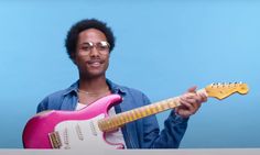 a man holding a pink and yellow guitar in his right hand while standing next to a blue wall