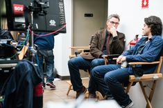 two men sitting on chairs in front of a camera and another man standing behind them