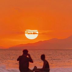 two people sitting on the beach watching the sun set