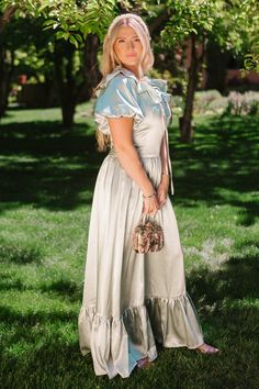 a woman standing in the grass holding a purse and wearing a dress with ruffles