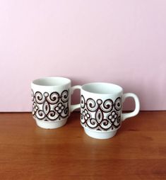 two coffee mugs sitting next to each other on top of a wooden table in front of a pink wall