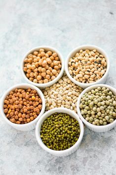 five white bowls filled with different types of food on a gray surface, including peas and lentils