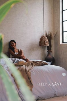 a woman sitting on top of a bed next to a plant in a room with white walls