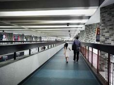 two people walking down a long walkway in a building with tiled walls and metal railings