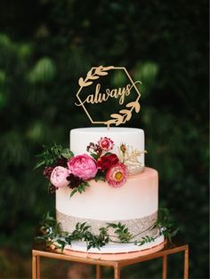 a white wedding cake with pink flowers and greenery on top, sitting on a gold stand