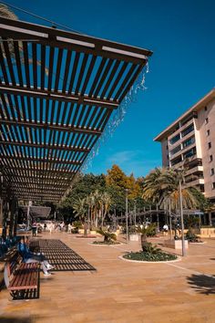 people are sitting on benches in the middle of an open area with palm trees and buildings behind them