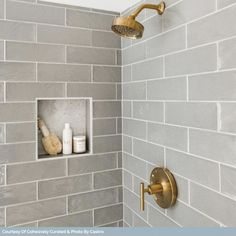 a bathroom with gray tile and gold faucet, shower head and hand held soap dispenser