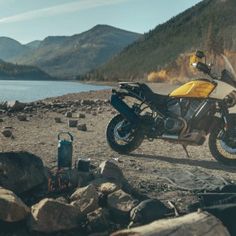 a yellow motorcycle parked on top of a dirt field next to a lake and mountains