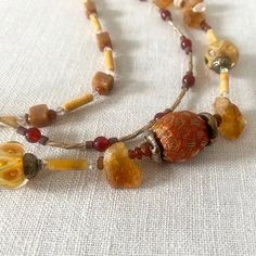 Beautiful, warm vintage beaded necklace with three strands combining a variety of gemstones and other beads. The necklace, dating to the late 1990s or early 2000s, has three strands of different lengths. The longest (and most ornate) has at its center a bead covered with gold and burnt orange fabric; the rest of the strand includes chunks of rough topaz, brown and white lampwork glass beads, slivers of baroque pearl, and small crystal beads. The next longest strand has round carnelian beads (about 5mm) with silver twist tube beads and small reddish-brown stone beads. And the shortest has square brown stone beads (agate or jasper?) mixed with long beige tubes (agate I think, but they could be a nice plastic) and small orange and crystal beads. The strands are securely strung and attached to Vintage Amber Beads Gems And Cabochons For Gift, Vintage Multi-strand Beaded Necklace With Colorful Beads, Vintage Multi-strand Beaded Necklaces With Colorful Beads, Vintage Multi-strand Colorful Beaded Necklaces, Vintage Amber Beaded Gems And Cabochons, Vintage Amber Beads, Gems, And Cabochons, Unique Amber Beaded Necklaces With Colorful Beads, Unique Amber Beaded Necklace With Colorful Beads, Vintage Amber Beads For Gifts