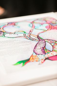 an open book with colorful designs on it sitting on a white table top next to a persons hand