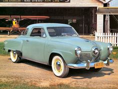 an old green car parked in front of a building with a plane on the roof