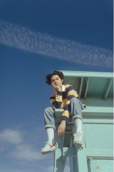 a young boy sitting on top of a metal ladder next to a blue building with white clouds in the background